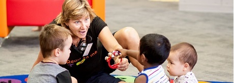 Library volunteer sits on colourful mat with two toddlers and one baby, one toddler reaches for the red musical bell shaker that the volunteer is holding out to him.