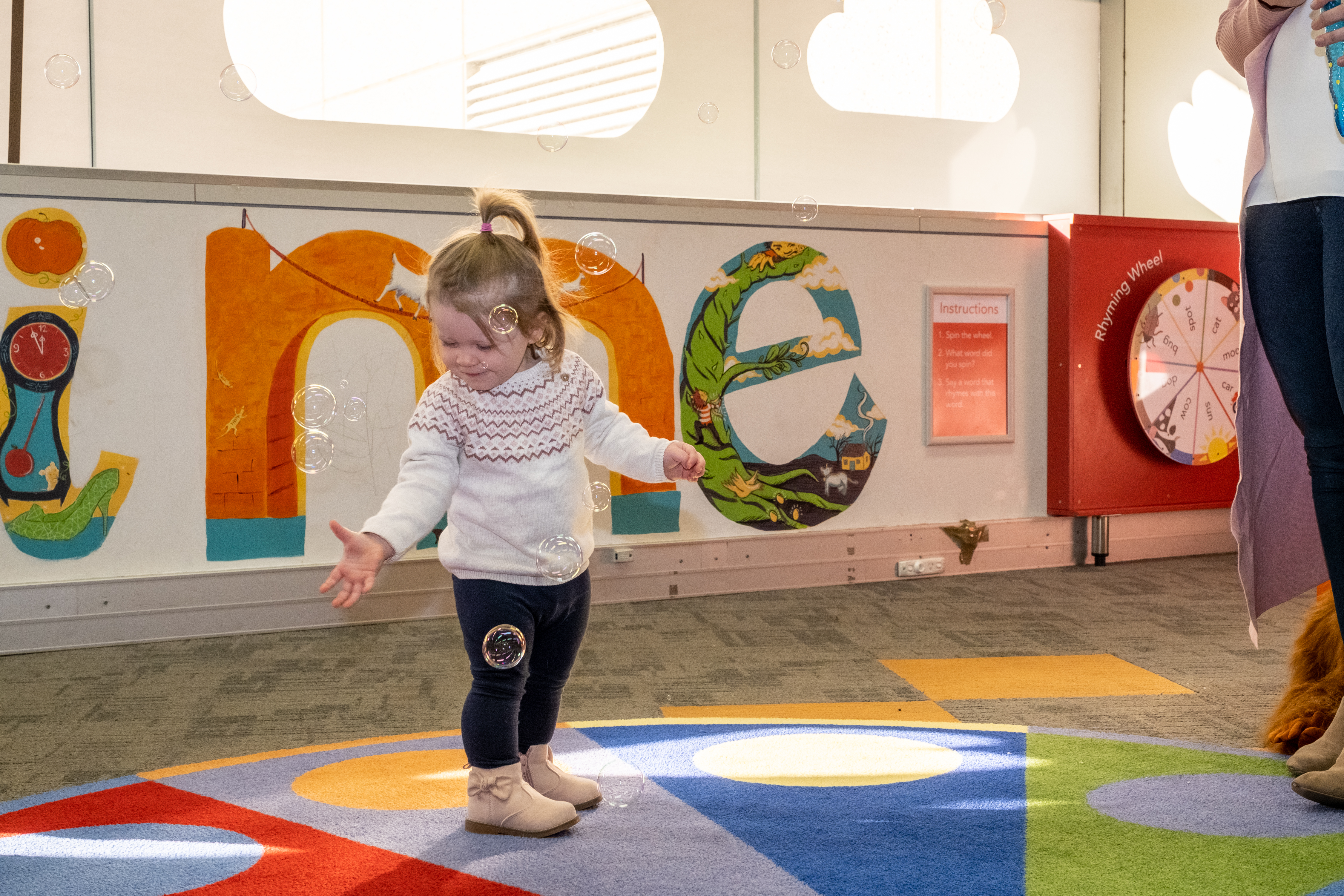 A toddler girl stands in a brightly lit library with Story Time painted on the walls, she is trying to catch bubbles.